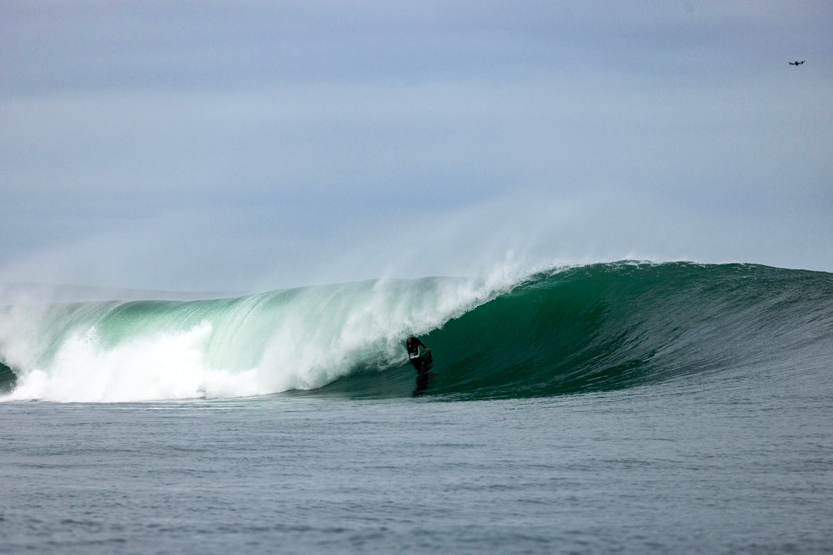 A surfer rides in XCEL wetsuit inside the barrel of a large green ocean wave under an overcast sky.