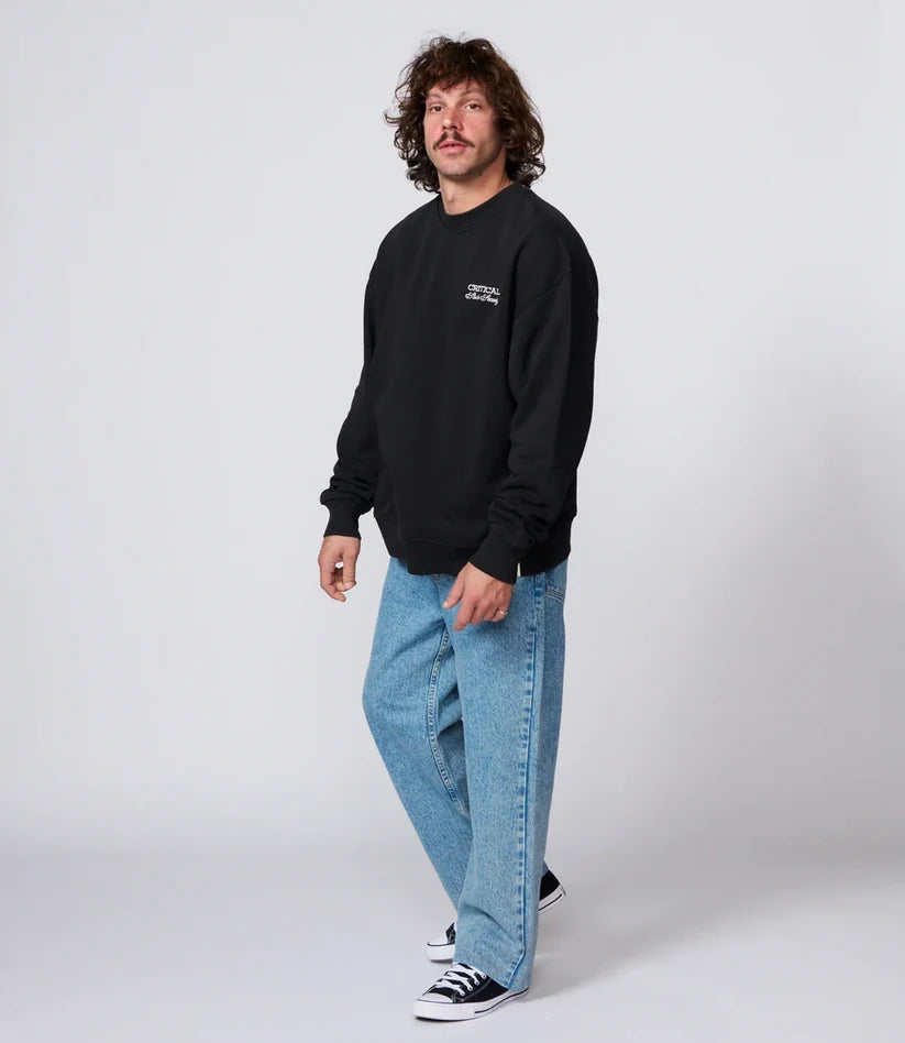 A man modeling a black crewneck sweatshirt with a small white graphic on the left chest, paired with light blue jeans and black-and-white sneakers, against a light gray studio background.