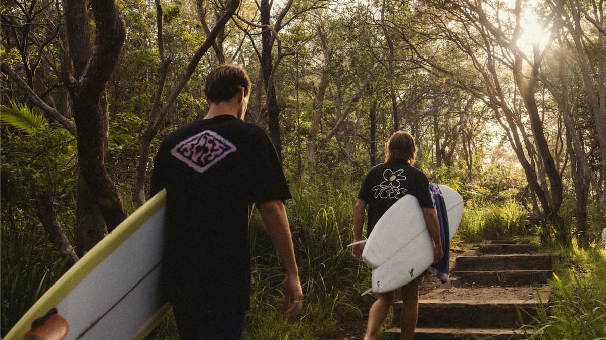 Two men holding surfboards walking through trees towards the sunlight