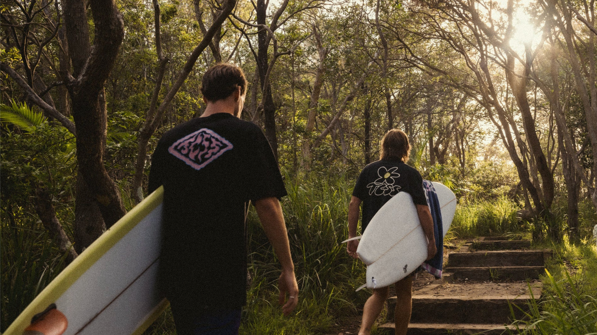 Two surfers walking up a wooded path carrying surfboards, seen from behind. Both wear black surf shirts with graphic logos on the back, with bright sunlight filtering through the trees.